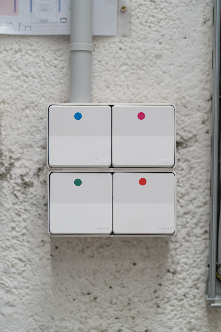 quatre interrupteurs sur un mur, chaque interrupteur a une gommette ronde d'une couleur différente, da haut en bas et de gauche à droite elles sont bleue, rose, verte et orange