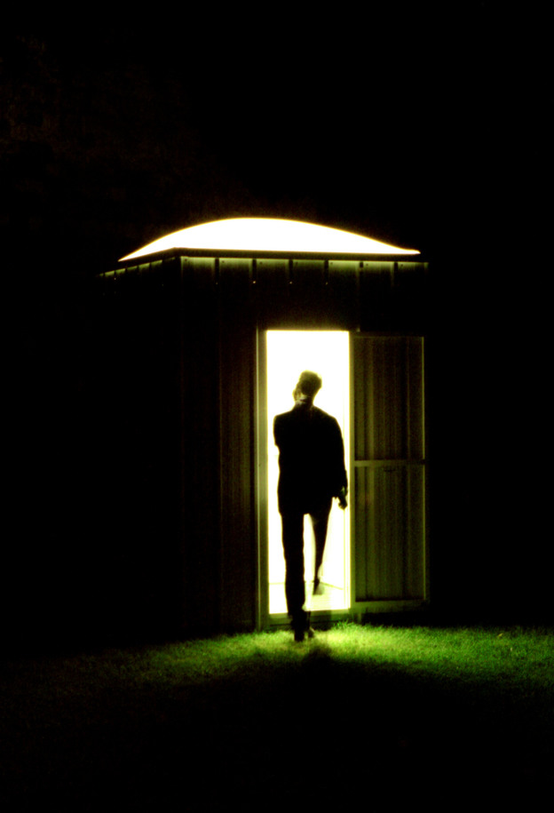 Dans la nuit noire un personnage vu de dos entre une cabane éclairée